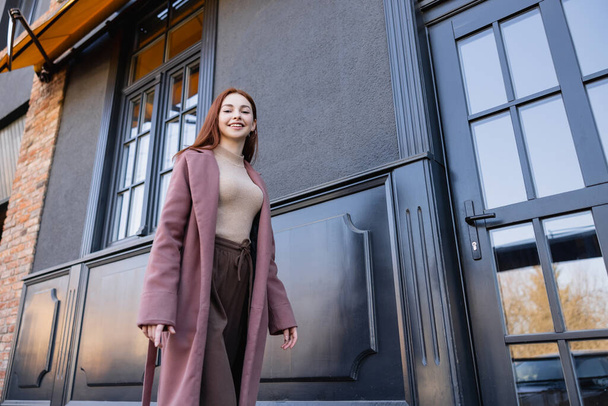 low angle view of young redhead woman in stylish coat smiling and walking near modern building  - Photo, Image