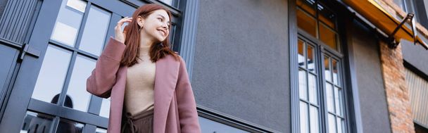 low angle view of happy woman in stylish coat adjusting red hair near modern building, banner - Foto, Imagen