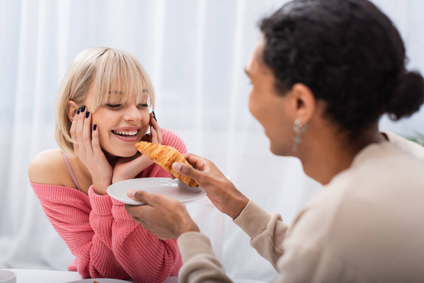 blurred african american man feeding blonde girlfriend with croissant  - Fotografie, Obrázek