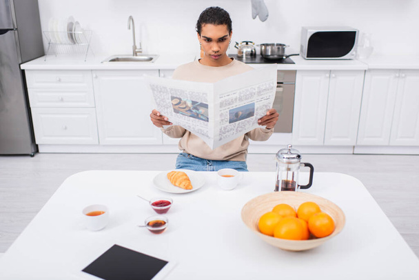 молодий афроамериканський чоловік читає газету біля сніданку та цифровий планшет на столі
 - Фото, зображення