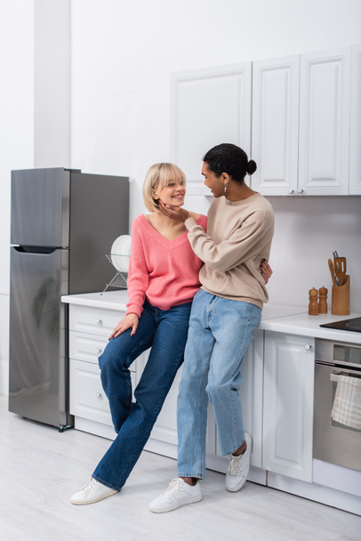 full length of smiling multiethnic couple looking at each other in modern kitchen  - Foto, imagen