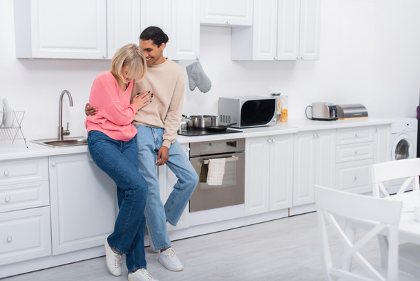 full length of pleased multiethnic couple standing in modern kitchen  - Foto, afbeelding