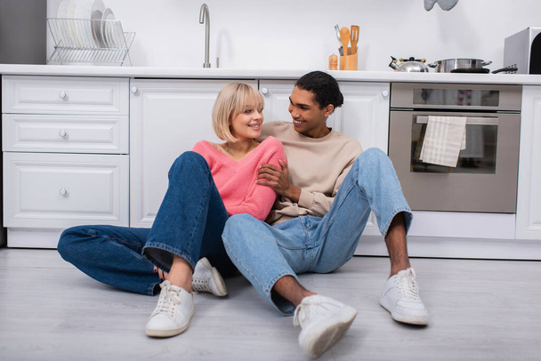 cheerful multiethnic couple sitting on floor in modern kitchen - Zdjęcie, obraz