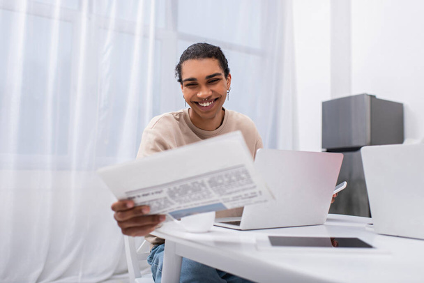 Ευτυχισμένος νεαρός Αφροαμερικάνος διαβάζει εφημερίδες κοντά σε gadgets  - Φωτογραφία, εικόνα