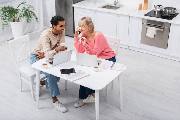high angle view of happy african american man looking at blonde girlfriend talking on smartphone while working from home - Photo, Image
