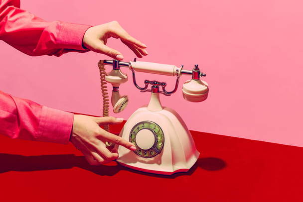 Pop art photography. Retro objects, gadgets. Female hand holding handset of vintage phone isolated on pink and red background. Vintage, retro fashion style - Zdjęcie, obraz