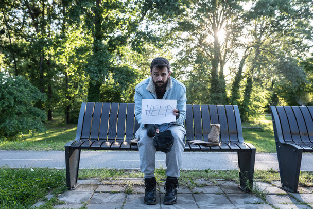 Mladý zoufalý nemocný bezdomovec se cítí opuštěný a smutný, když sedí venku. Nemocný muž bez práce potřebuje pomoc. Koncept sociální problematiky bezdomovectví. - Fotografie, Obrázek