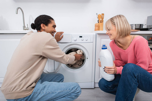 щаслива блондинка тримає пляшку з миючим засобом біля афроамериканського чоловіка, поклавши одяг у пральну машину
 - Фото, зображення