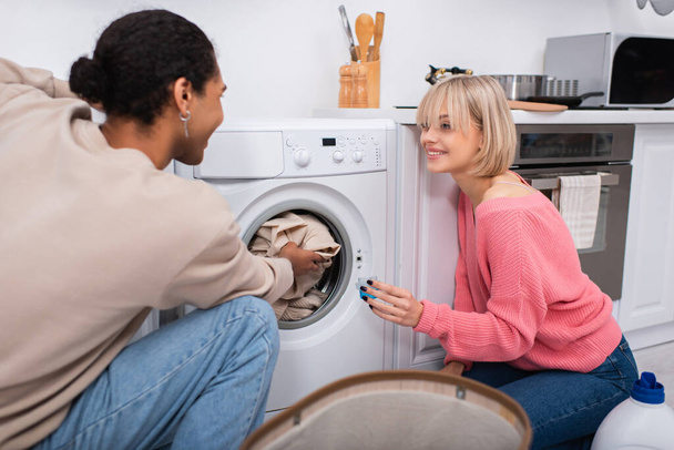 allegra donna bionda in possesso di tazza con detergente vicino all'uomo americano africano che mette i vestiti in lavatrice - Foto, immagini