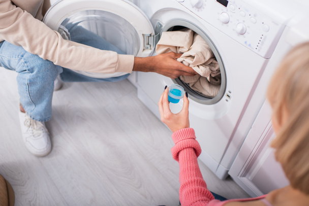 przycięty widok kobiety trzyma filiżankę z detergentem w pobliżu Afryki amerykański mężczyzna wprowadzenie ubrania w pralce - Zdjęcie, obraz