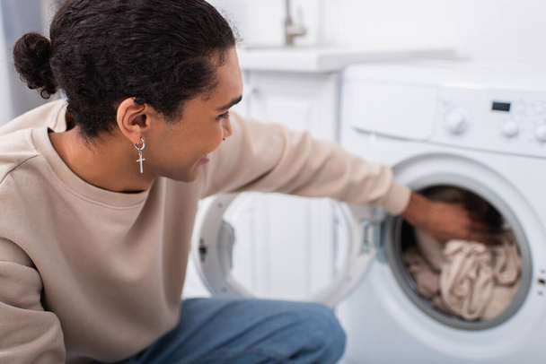 Mutlu Afrikalı Amerikalı adam çamaşır makinesine çamaşır koyuyor. - Fotoğraf, Görsel