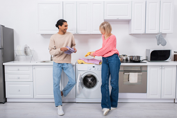cheerful interracial couple holding clean and colorful clothes  - Photo, Image