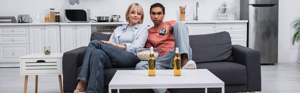 african american man holding remote controller and clicking channels near blonde girlfriend in living room, banner - Fotó, kép