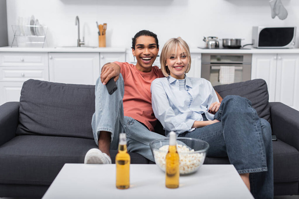 happy interracial couple watching movie near beer bottle and popcorn on coffee table - 写真・画像