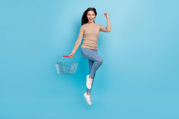 Photo of astonished lady jump hold supermarket basket raise hand wear beige sweater isolated blue color background - Zdjęcie, obraz