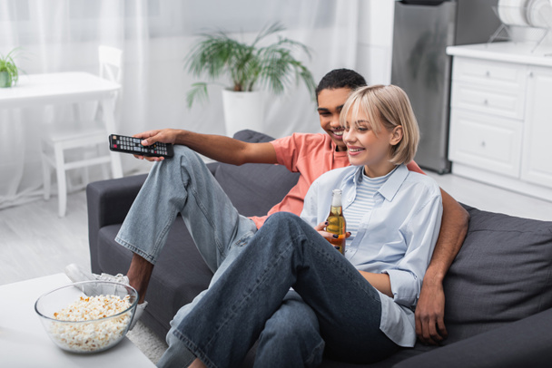 happy african american man looking at cheerful woman with bottle of beer while watching movie - Photo, Image