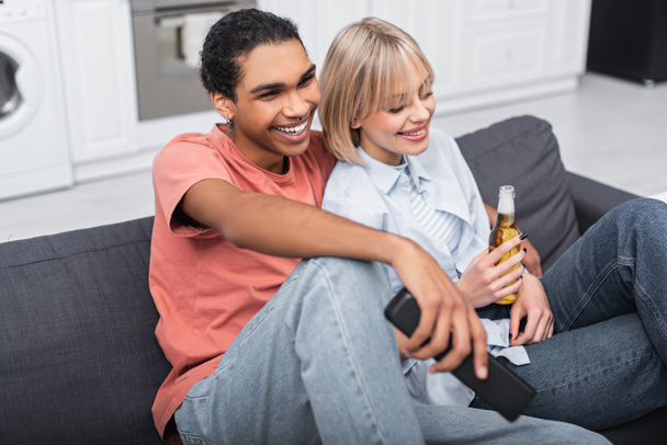 happy blonde woman holding bottle of beer near smiling african american man with remote controller  - Foto, Imagem