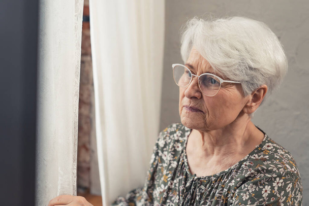 bothered upset grey-haired grandmother wearing glasses and peeking through the window - Фото, изображение
