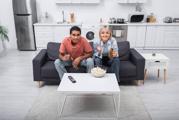 érzelmi interracial pár éljenzés és a kezében üveg sör, miközben nézi sport mérkőzés a nappaliban  - Fotó, kép