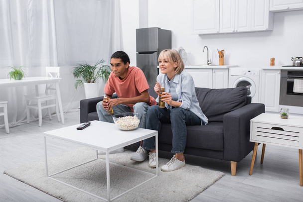 young multiethnic couple holding bottles with beer while watching sport match in living room  - Φωτογραφία, εικόνα
