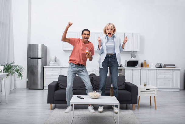 happy multiethnic couple jumping while watching sport match in living room  - Φωτογραφία, εικόνα