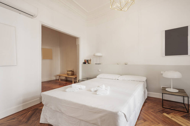Schlafzimmer mit Doppelbett, weißen Kissen, Kommode im Hintergrund, weißen Nachttischen und Bettwäsche, grauem Kopfteil aus Mauerwerk und rötlichem Holzboden - Foto, Bild