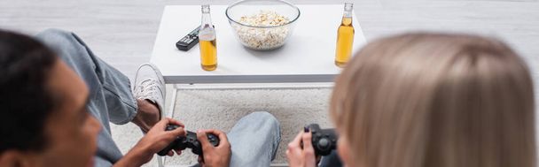 KYIV, UKRAINE - DECEMBER 6, 2021: popcorn and beer near multiethnic couple playing video game, banner - Foto, afbeelding