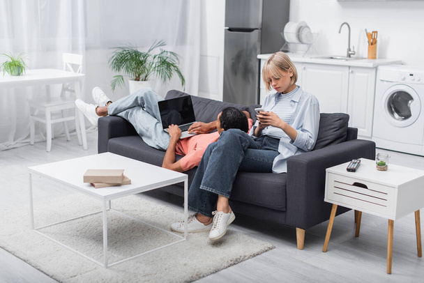 african american man using laptop with blank screen and lying near blonde girlfriend with cellphone on couch - Valokuva, kuva