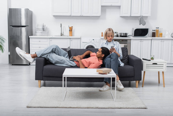 african american man using laptop with blank screen and lying near blonde girlfriend with smartphone on couch - Φωτογραφία, εικόνα