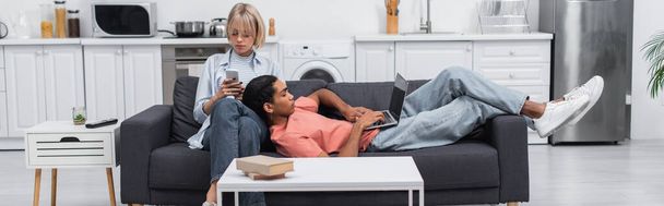 african american man using laptop with blank screen and lying near blonde girlfriend with smartphone on couch, banner - Valokuva, kuva