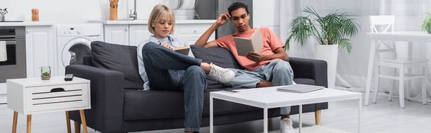 young african american man and blonde woman reading books near gadgets on coffee table, banner - Foto, Imagem