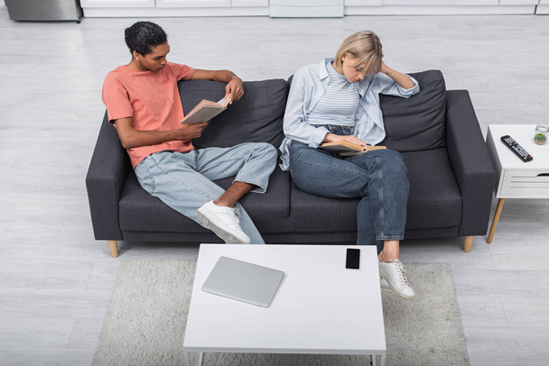 top view of young african american man and blonde woman reading books near gadgets in living room  - Foto, immagini