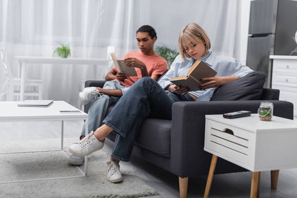 young blonde woman and african american man reading books near gadgets in modern living room  - Foto, afbeelding