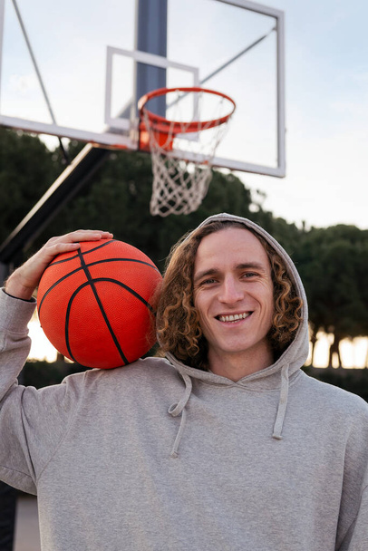vertical portrait of a smiling young man holding a basketball on a city court, concept of urban sport in the street, copy space for text - Фото, зображення