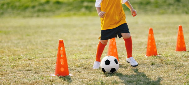 Dziecko poprawy umiejętności piłki nożnej uruchomiony piłkę na stożki wiertarki. Młody chłopiec z piłką nożną na treningu nosi korki piłkarskie - Zdjęcie, obraz