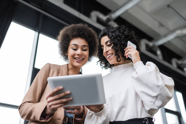 Vue en angle bas de souriantes femmes d'affaires interraciales utilisant une tablette numérique et parlant sur smartphone au bureau  - Photo, image