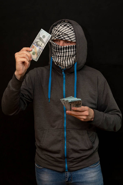 A man in a hoodie looks at a $100 bill, his face is covered with an Arab scarf, dark background, selective focus. - Zdjęcie, obraz
