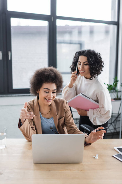 Empresarias interracial positivas usando laptop y hablando en smartphone en la oficina  - Foto, imagen