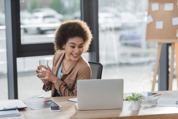 Cheerful african american businesswoman holding glass of water near laptop and documents in office  - Foto, Imagen