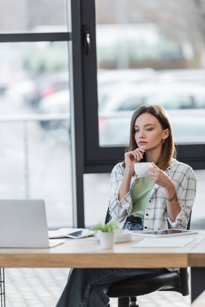 Pensive businesswoman holding cup near blurred laptop in office  - Photo, Image