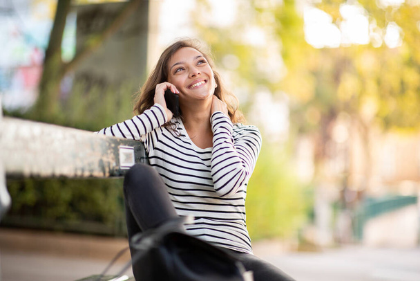 Portret van een lachende jonge vrouw die op een bankje in het park zit te praten met een mobiele telefoon - Foto, afbeelding