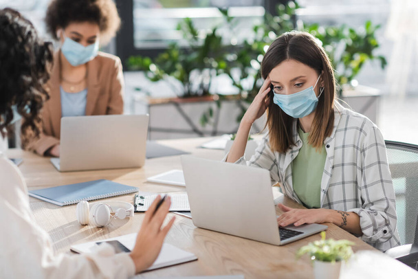 Businesswoman in medical mask using laptop near blurred interracial colleagues in office  - Foto, afbeelding