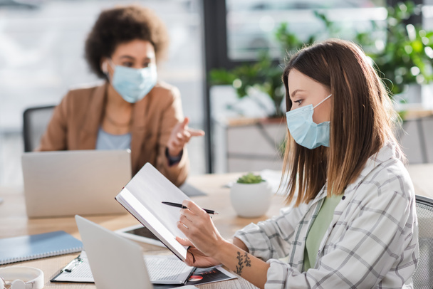 Businesswoman in medical mask holding notebook near blurred laptop and african american colleague in office  - Fotó, kép