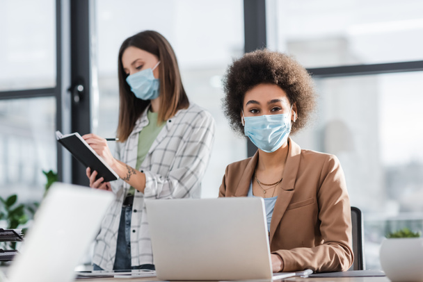 African american businesswoman in medical mask looking at camera near laptop and colleague in office  - Foto, Imagem