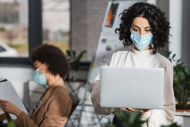 Muslim businesswoman in medical mask using laptop near blurred african american colleague in office  - Foto, imagen