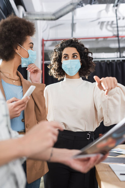Arabian businesswoman in medical mask pointing at blurred paper near interracial colleagues in office  - Foto, Imagem