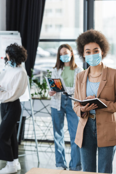 African american businesswoman in medical mask holding notebook while working in office  - Photo, Image