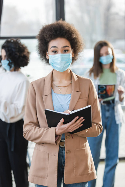 Young african american businesswoman in protective mask holding notebook and looking at camera in office  - Foto, immagini