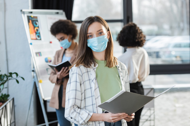 Young businesswoman in medical mask holding paper folder near blurred multiethnic colleagues  - Fotoğraf, Görsel