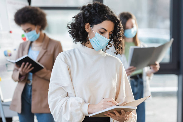 Arabian businesswoman in medical mask writing on notebook near blurred colleagues in office  - Photo, image